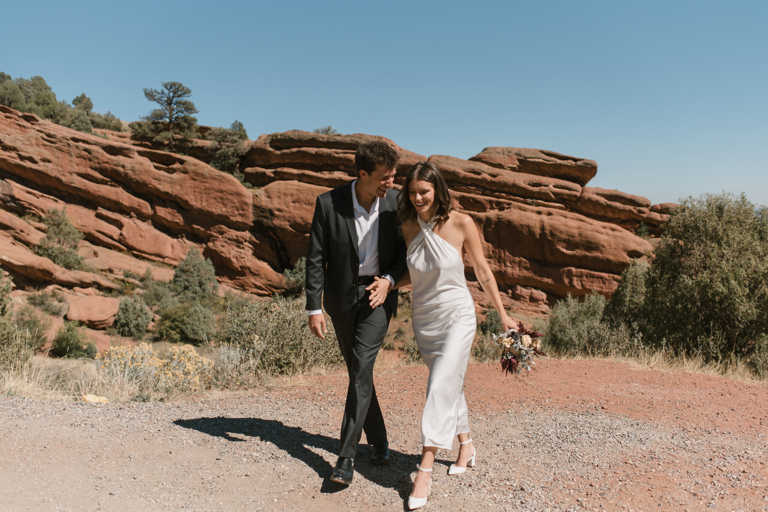 bridal couple holding hands and walking with red rocks behind them 