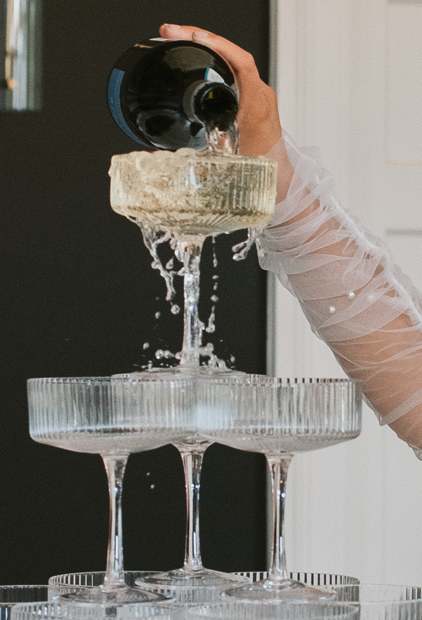 close up of bride's arm with wedding dress sleeve, pouring champagne onto tower