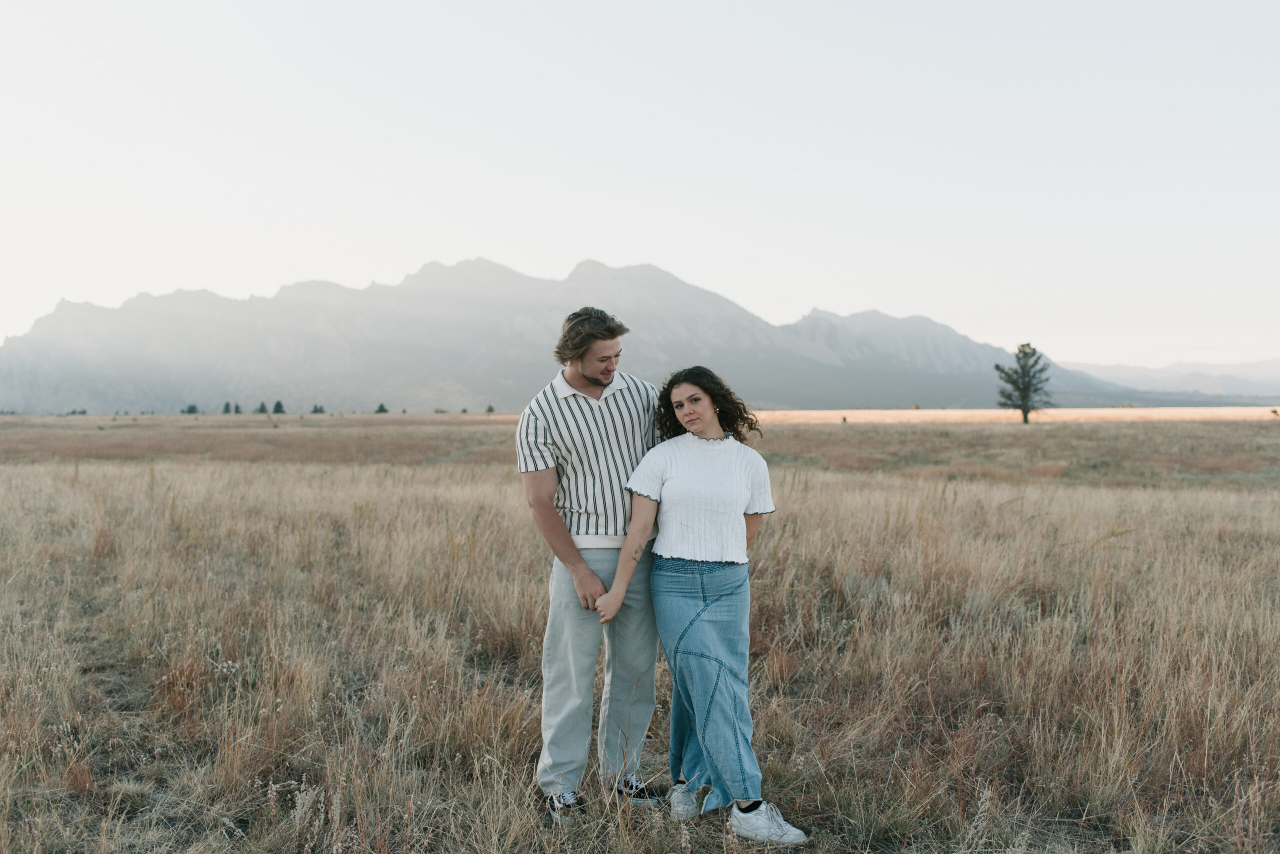 woman looking at the camera and man looking at her as they stand in a field during a couples photoshoot 