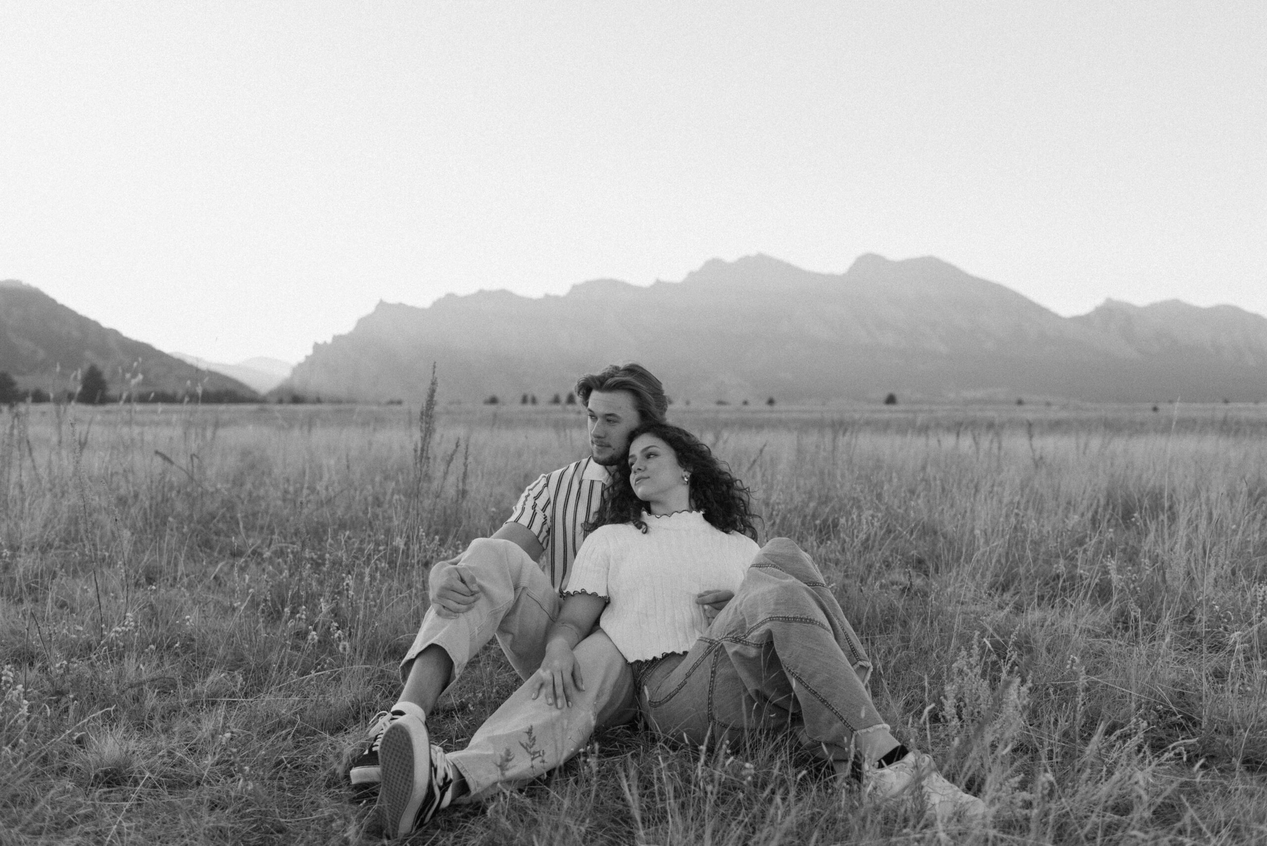 couple sitting in the grass and woman leaning her back against man's chest as they both look to the side