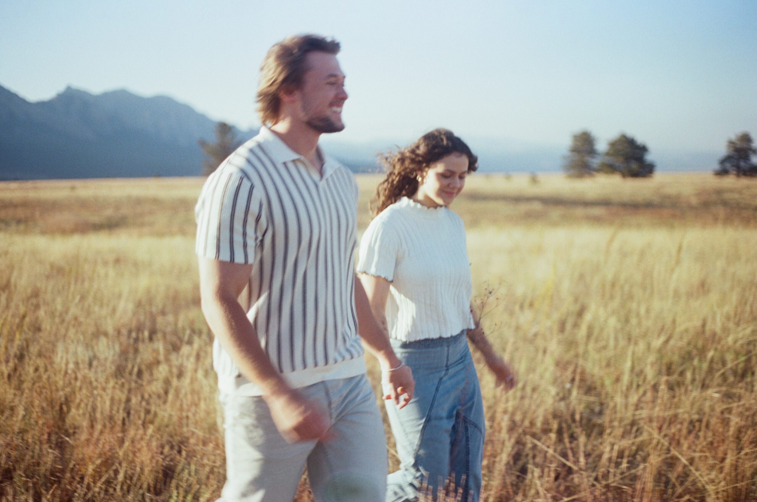 couple holding hands, smiling and walking through a open grass field 