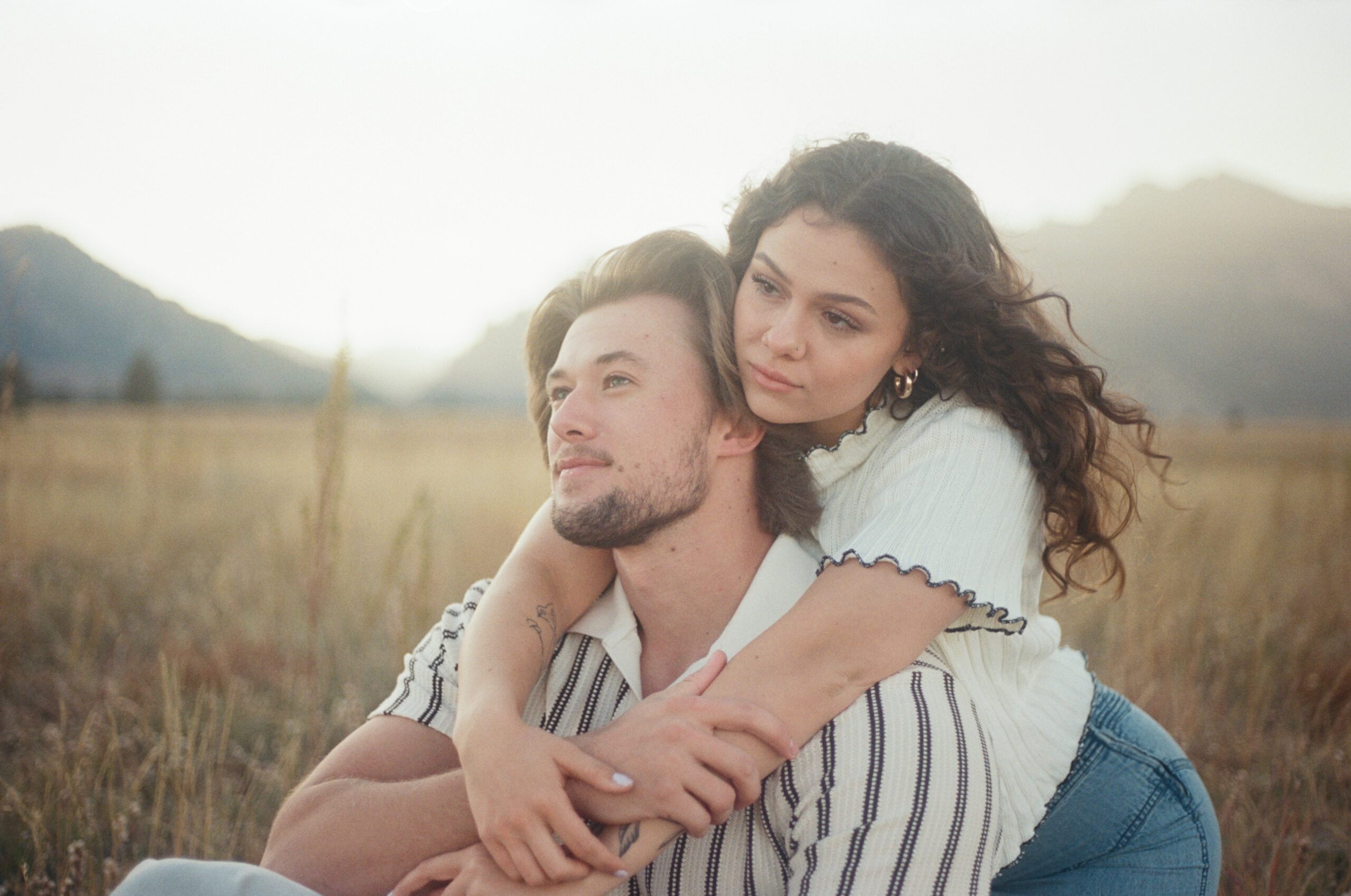 film photography of a woman holding her boyfriend from behind as they sit in a grass field 