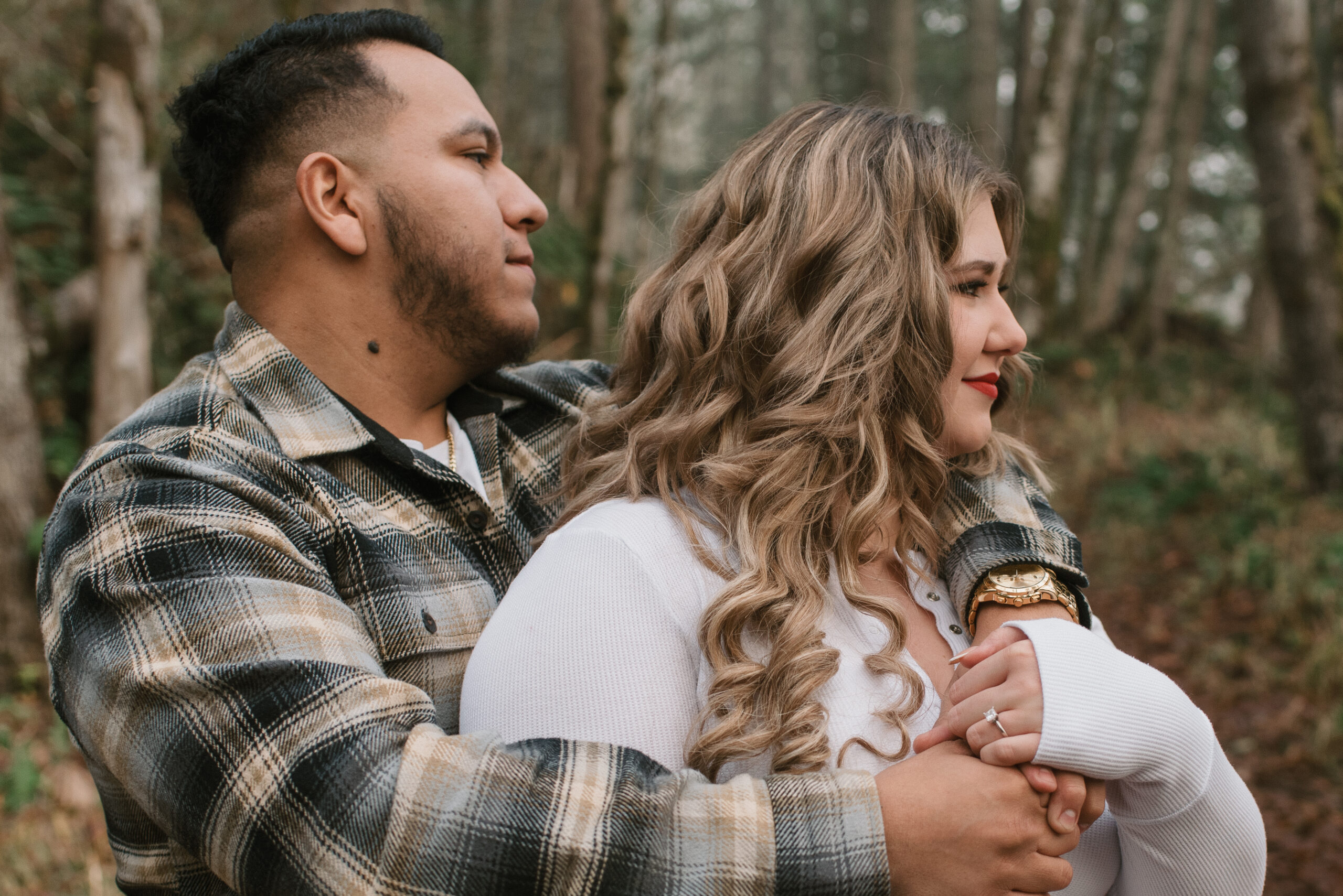 fiance holding other fiance from behind as they look off into the forest 