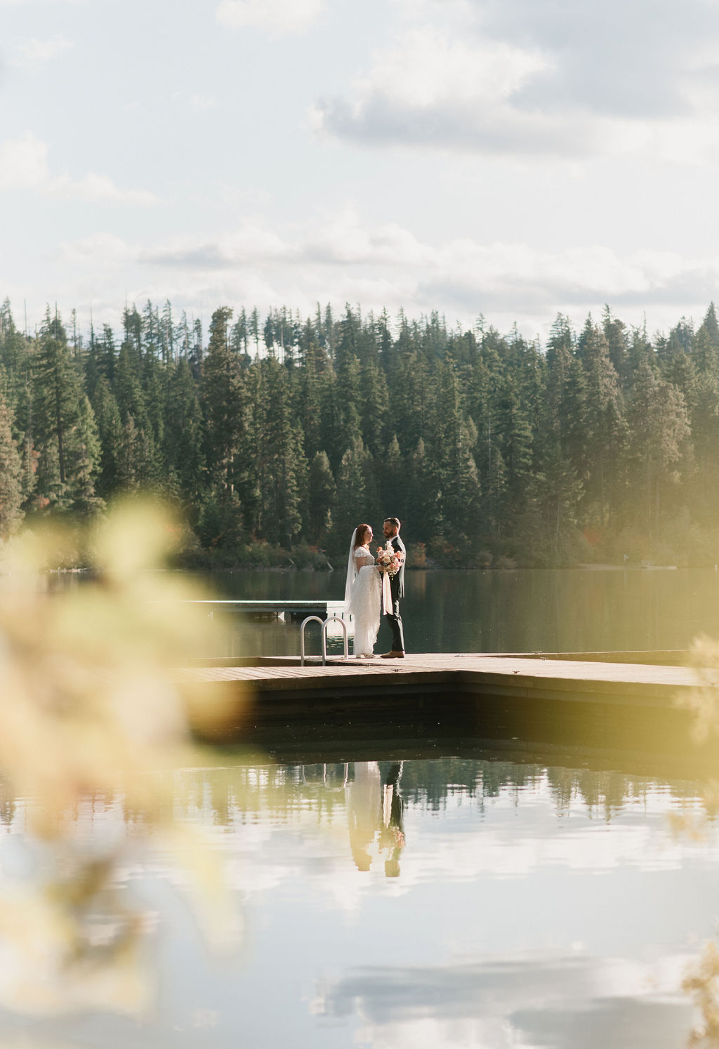 bride and groom standing on the dock together with trees and the lake in the background 