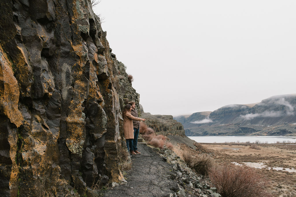 couple on a trail next to a rock wall looking out at the frozen lake during their modern engagement pictures