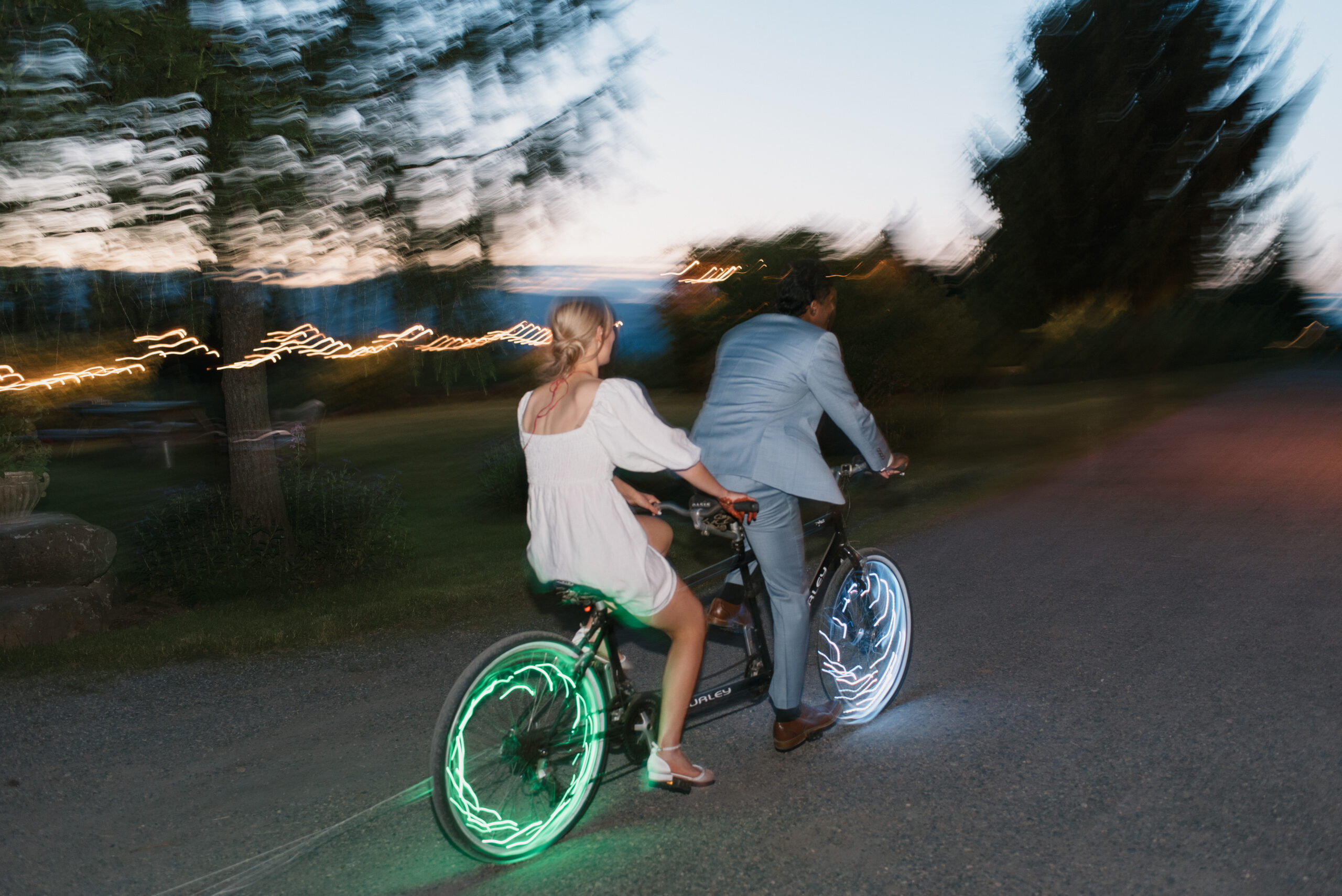 a slow shutter photo of the bride and groom on a tandem bike after sunset