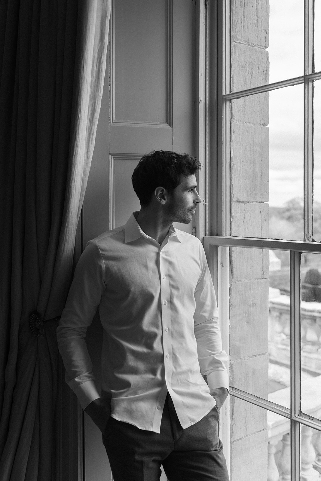 Groom buttoning shirt near tall window with soft Irish light, Hilton Park Manor wedding morning