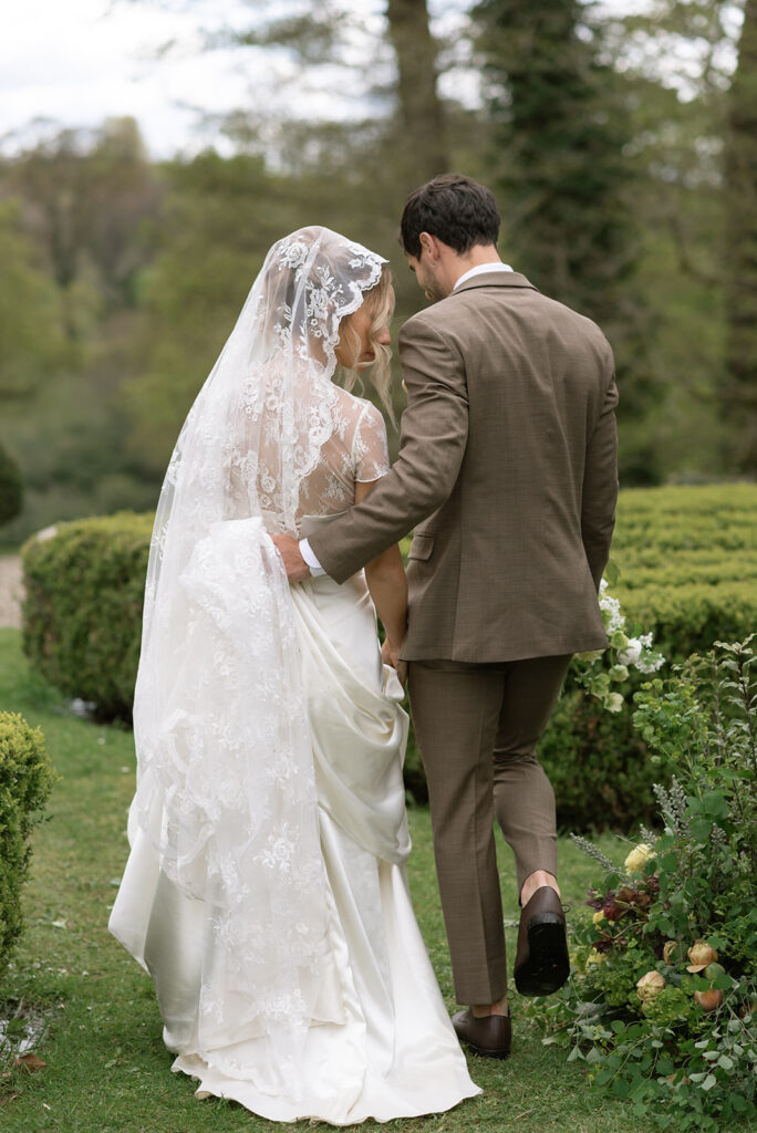 bride and groom walking together as groom holds wedding dress in lush gardens in ireland